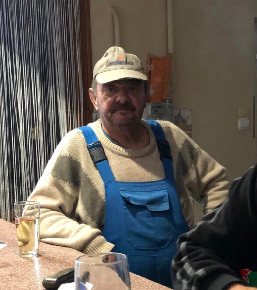 Ospalý muž v modrých montérkach a béžovom svetri stojí pri bare s pohárom piva.