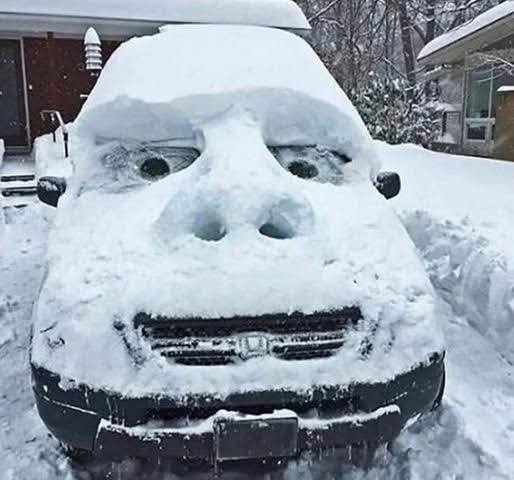 Auto pod snehom s vyrezanou tvárou, vytvára vtipný výraz v zimnom prostredí.