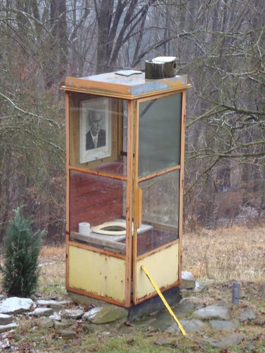 Starý telefónny automat prerobený na vonkajšie WC v lese, s fotografiou na stene.