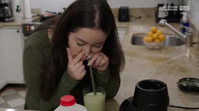 Osoba v kuchyni inhaluje zelený nápoj cez slamku, okolo sú mlieko a mixér.