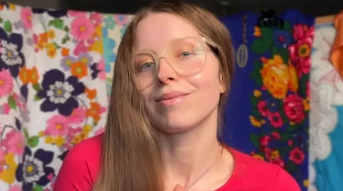 Žena s okuliarmi a dlhými vlasmi pred farebným kvetinovým pozadím v ružovom tričku.