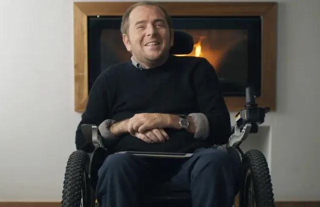 Osoba na vozíku v interiéri, s krbom v pozadí, usmieva sa.