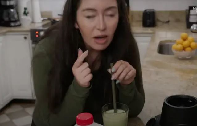 Žena v kuchyni ochutnáva zelené smoothie s vyjadrením prekvapenia. Na pozadí sú spotrebiče a misa s citrónmi.