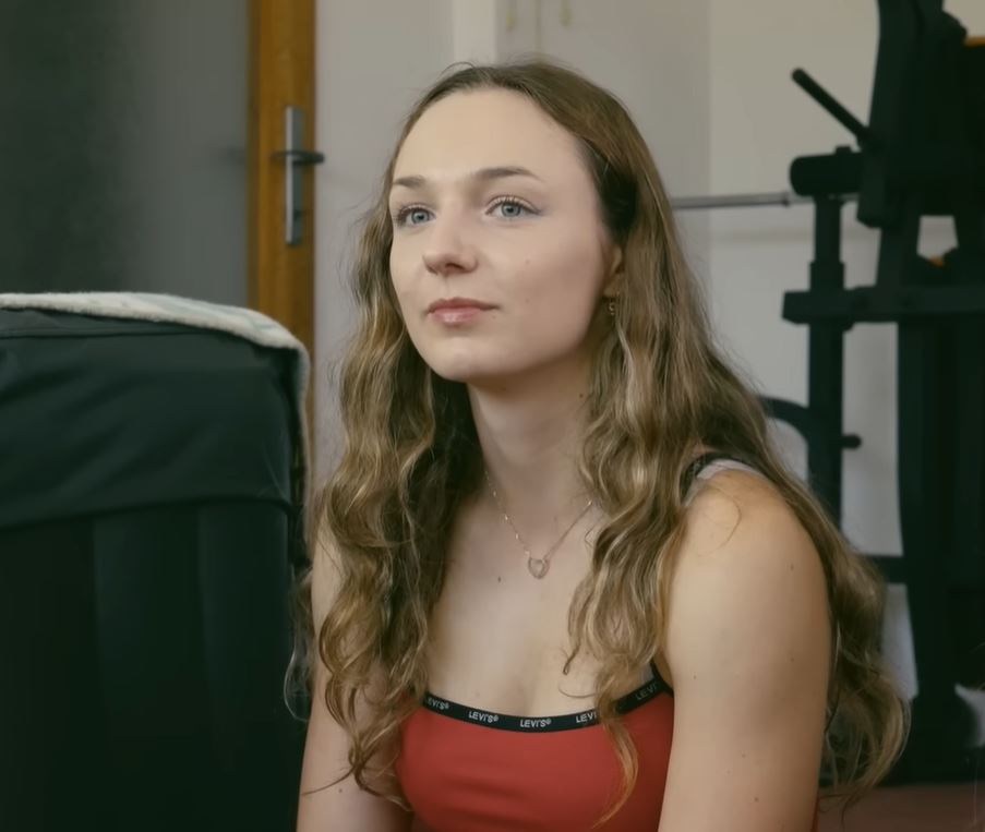 Mladá žena s dlhými vlasmi v červenom topu sedí v interiéri blízko cvičebných zariadení.