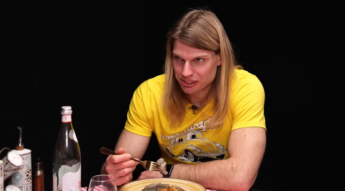 Muž v žltom tričku v reštaurácii, drží vidličku nad tanierom s jedlom, na stole fľaša s vodou.