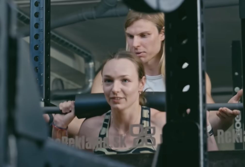 Žena so vzpieračskou činkou v posilňovni, zatiaľ čo ju muž povzbudzuje.