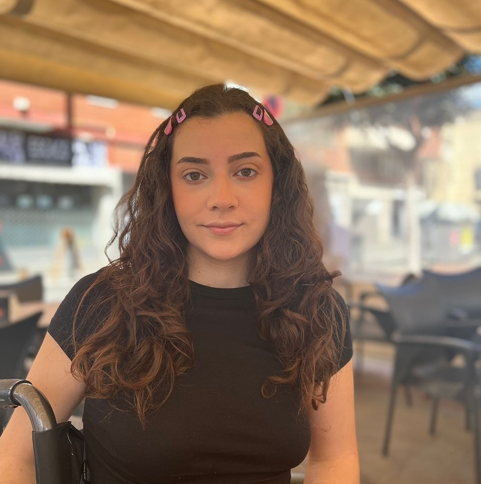 Žena s dlhými kučeravými vlasmi a čiernym tričkom sedí vonkajšej kaviarni, usmieva sa a pozerá do fotoaparátu.