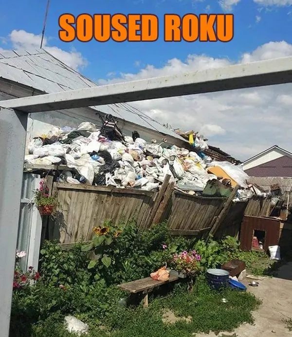 Dom so záhradou a hromadou odpadu za plotom, nápis Soused roku.