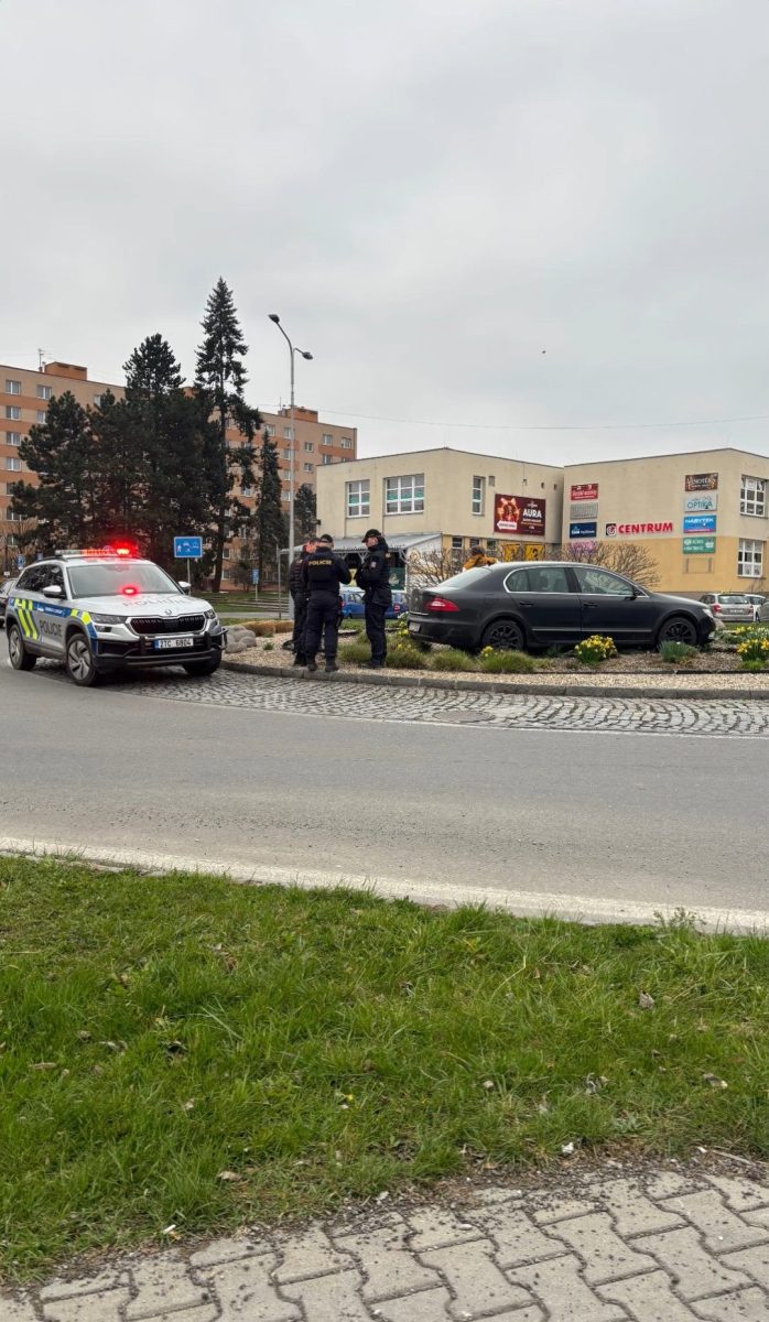 Policajti riešia situáciu s autom na kruhovom objazde pred budovou. V pozadí sú viacposchodové budovy a zeleň.