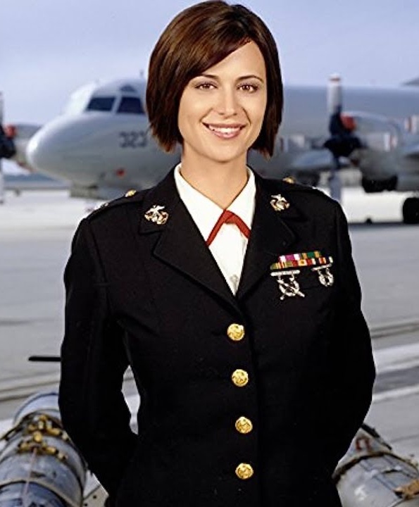 Žena v uniforme námorného letectva stojí pred vojenským lietadlom na letisku.