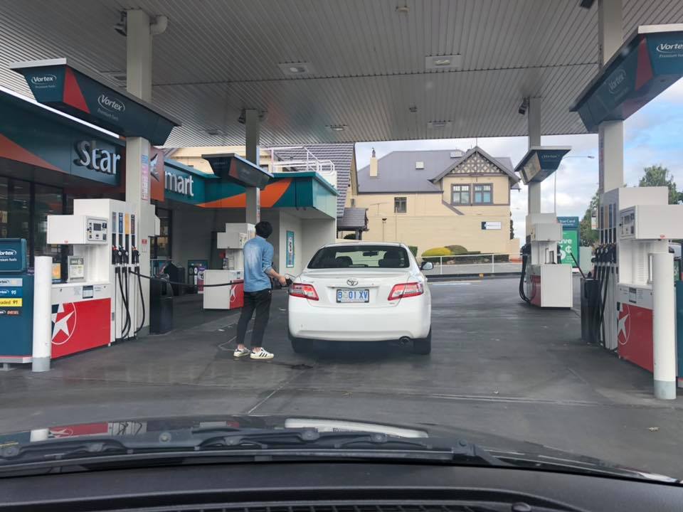 Auto na benzínovej stanici, muž tankuje palivo. Pozadie ukazuje obchod a budovy.