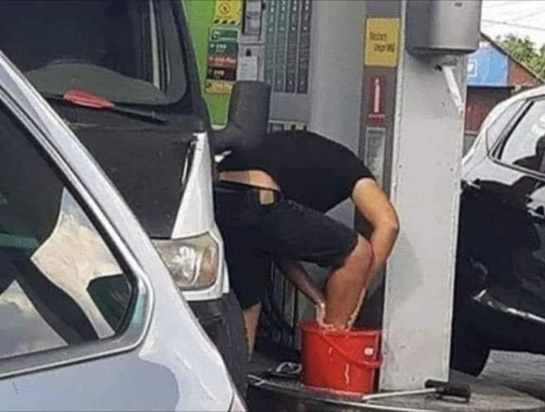 Osoba umýva nohy v červenom vedre na benzínovej pumpe vedľa auta.