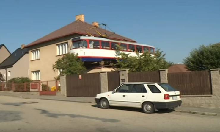 Dom s električkou na streche, zaparkované auto pred bránou, modrá obloha. Netypická scenéria na ulici.