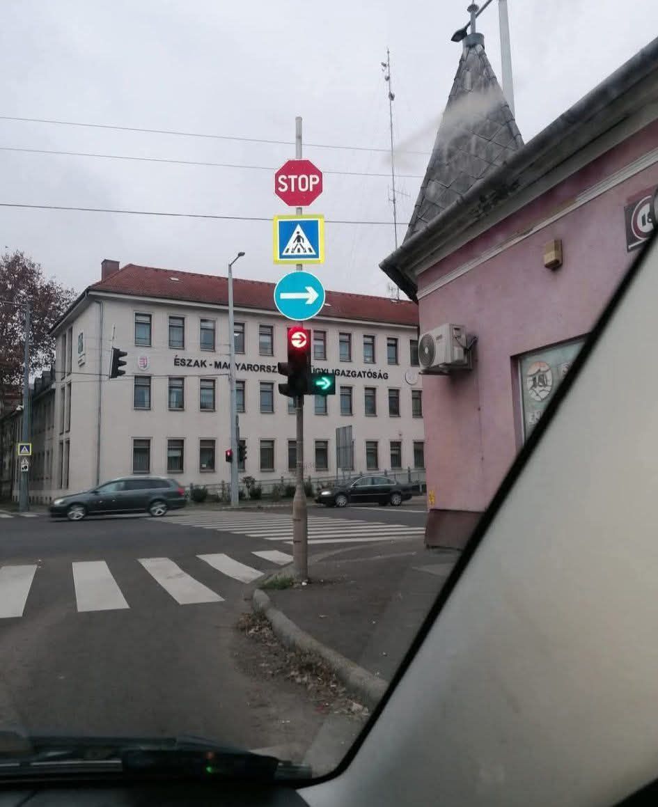 Dopravné značenie so semaforom na križovatke pri chodníku, za ním budova s nápisom v maďarčine.