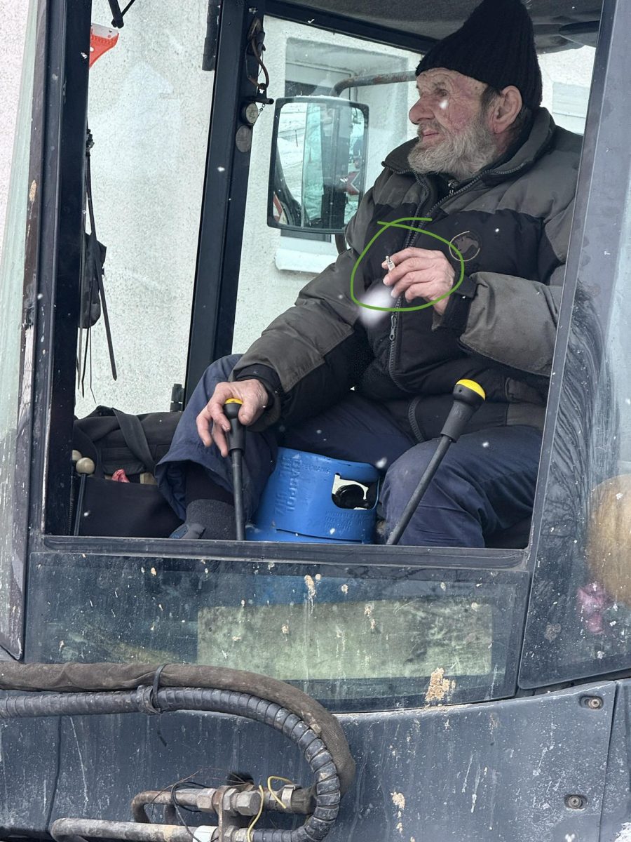 Muž v kabíne traktora, oblečený v zimnom oblečení, drží cigaretu.