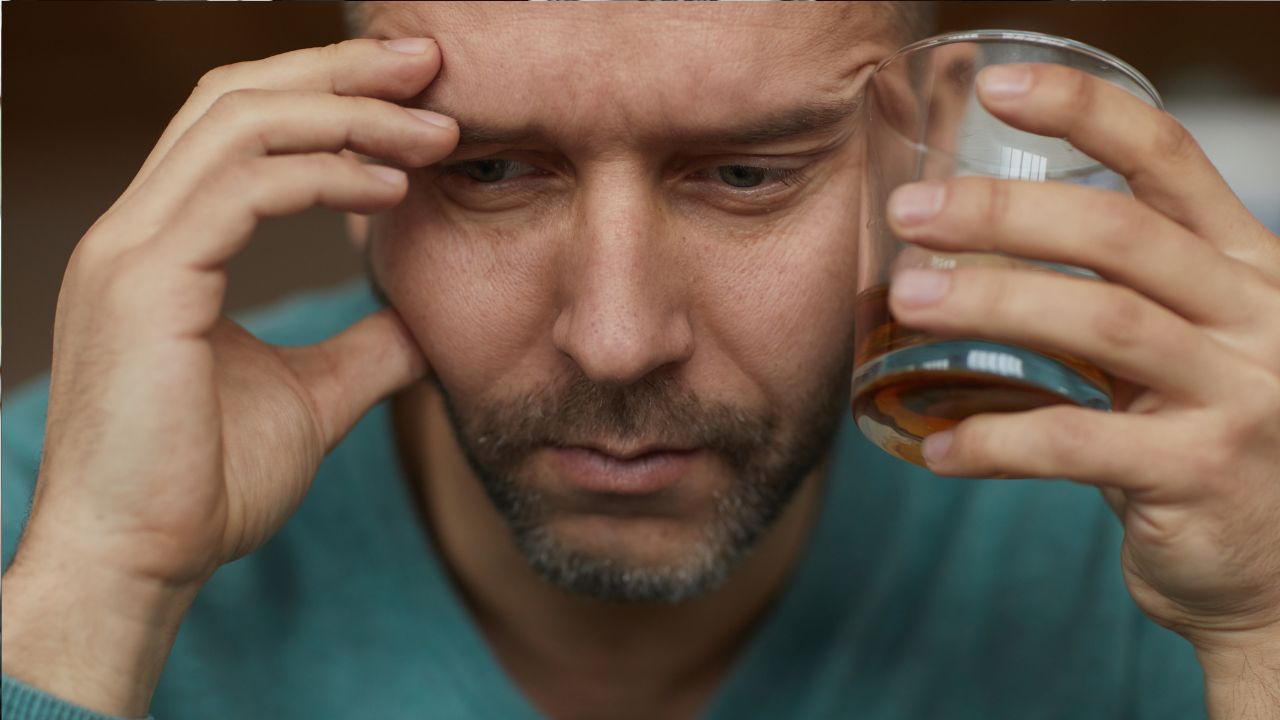 Muž v modrom svetri, zamyslene drží pohár whisky pri hlave, výraz sústredenia a zamyslenia na tvári.
