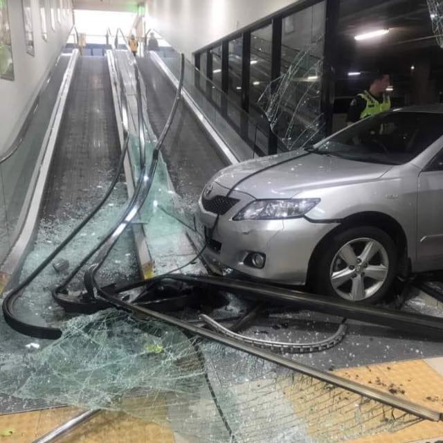 Auto havarovalo na eskalátore, rozbité sklo a kovové zábradlie. Policajt na mieste nehody v obchodnom centre.