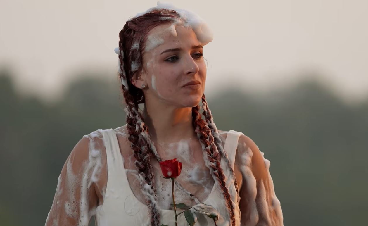 Žena s vrkočmi pokrytá penou drží červenú ružu, prírodné pozadie, dramatická atmosféra.
