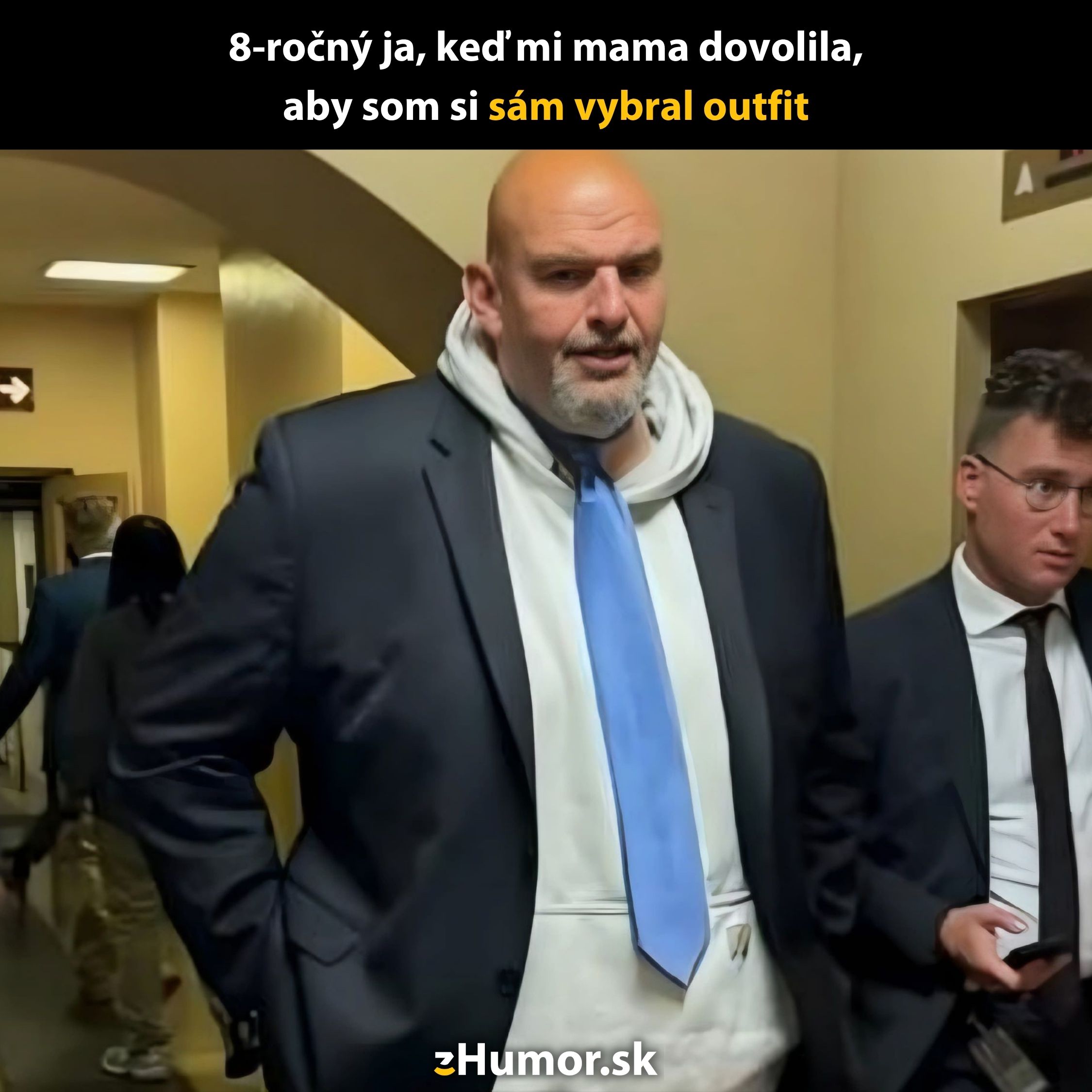 Muž v saku a mikine s kapucňou na chodbe; vedľa neho stojí iný muž v obleku.