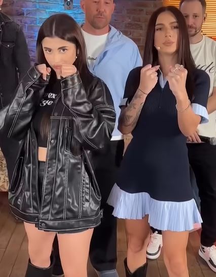 Dve ženy v modernom oblečení pózujú pred kamerou, s vážnymi výrazmi a gestami rukami v interiéri.