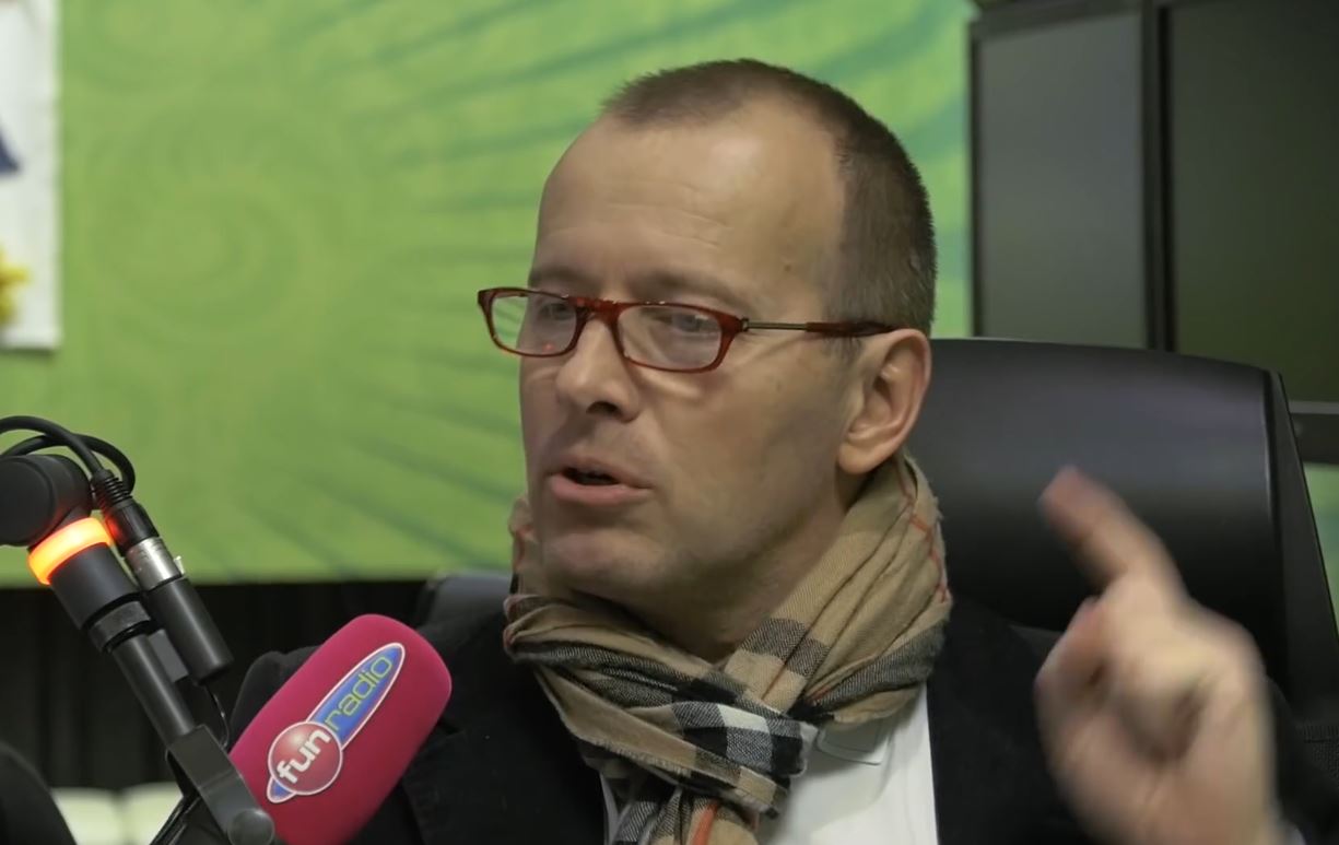 Osoba v rádiovom štúdiu pri mikrofóne diskutuje; má okuliare a šál, zelené pozadie.