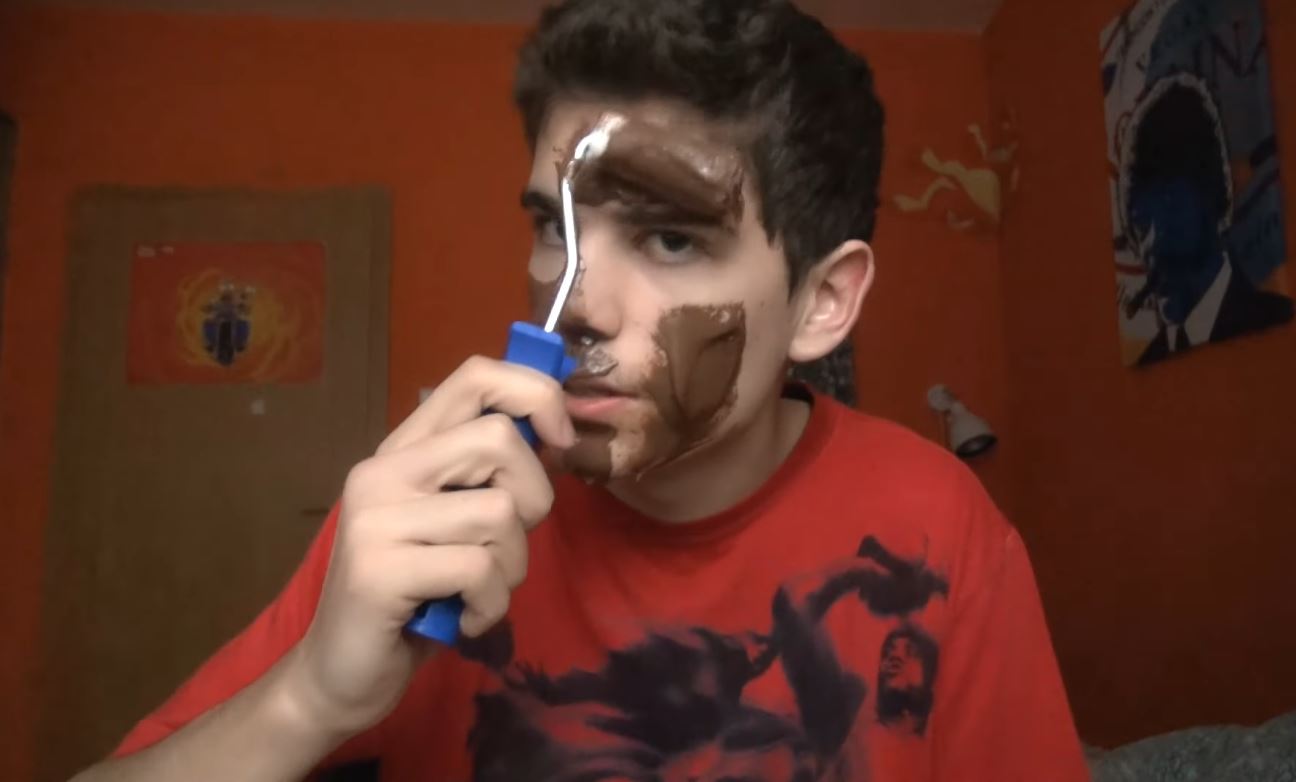 Muž si nanáša čokoládovú masku na tvár pomocou maliarskej valčekovej kefy v izbe s oranžovými stenami.