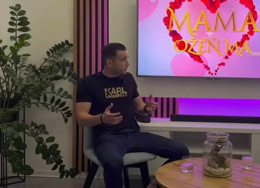 Muž diskutuje v show Mama, ožeň ma, sedí na stoličke pri obrazovke so srdcovým logom.
