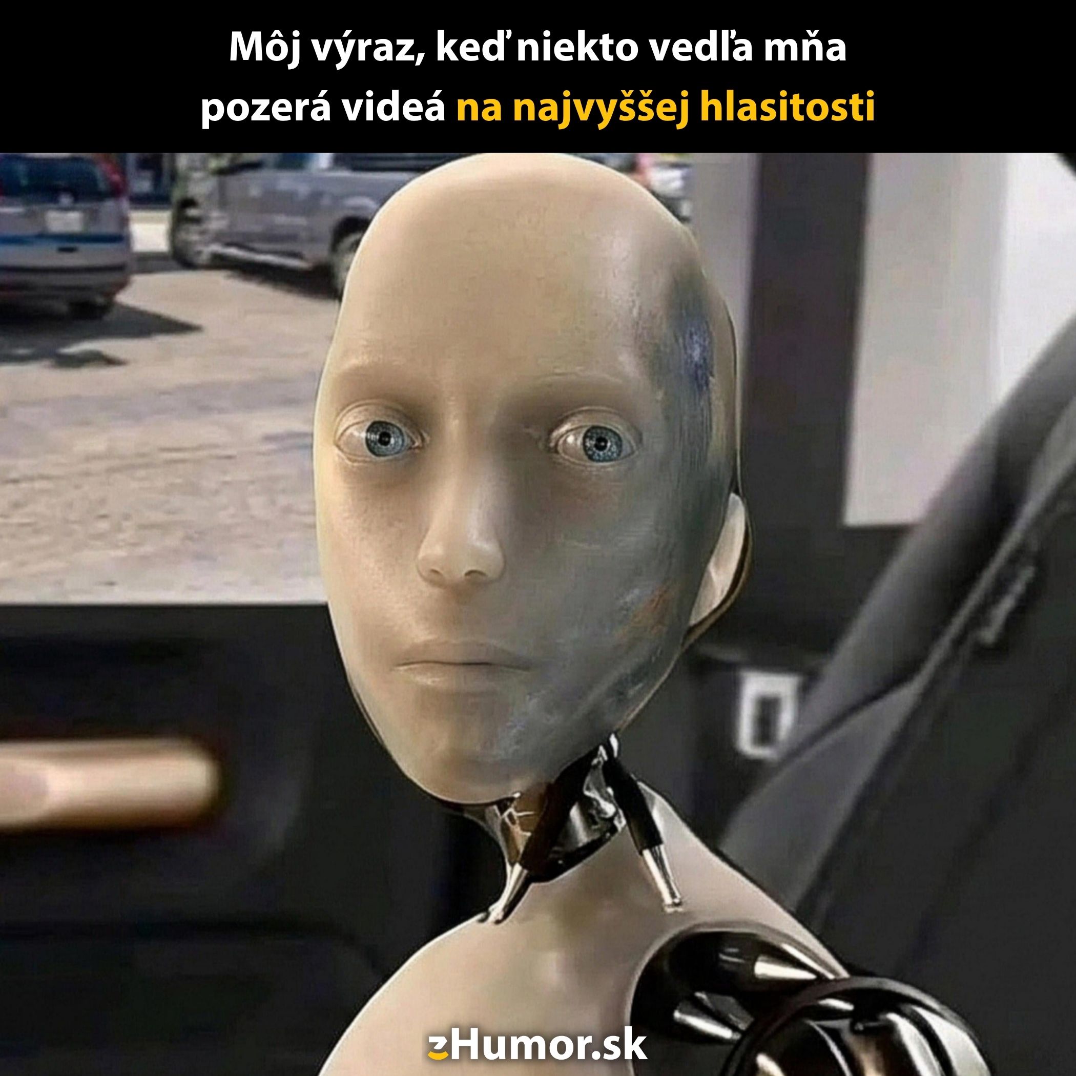 Robot s neutrálnym výrazom sedí v aute, fotka s vtipným textom o hlasitých videách.