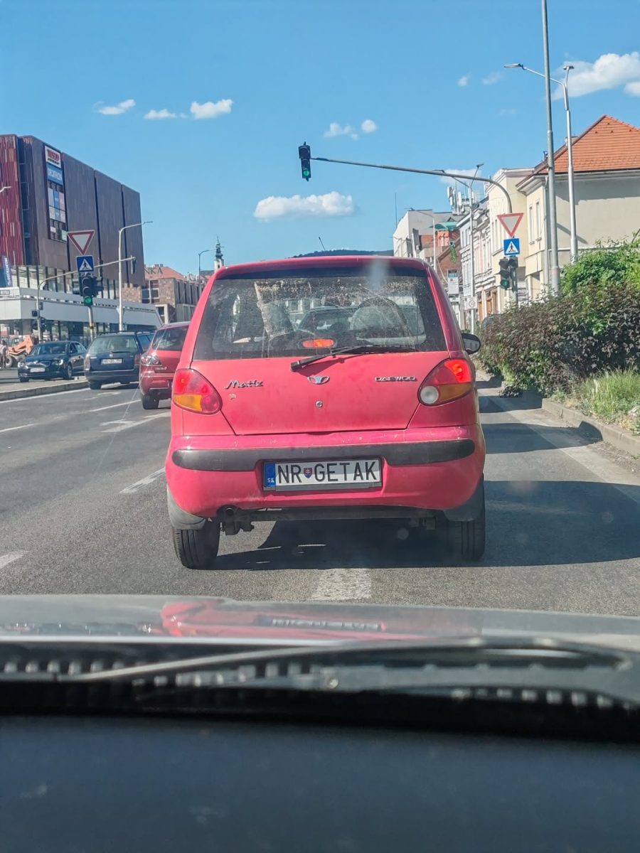 Malé červené auto s poznávacou značkou, jazda mestom za slnečného počasia, modrá obloha a dopravné svetlá.