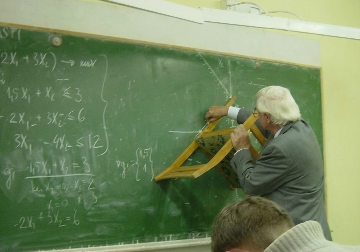 Učiteľ vysvetľuje matematiku pri tabuli, pomáhajúc si stoličkou ako pomôckou.