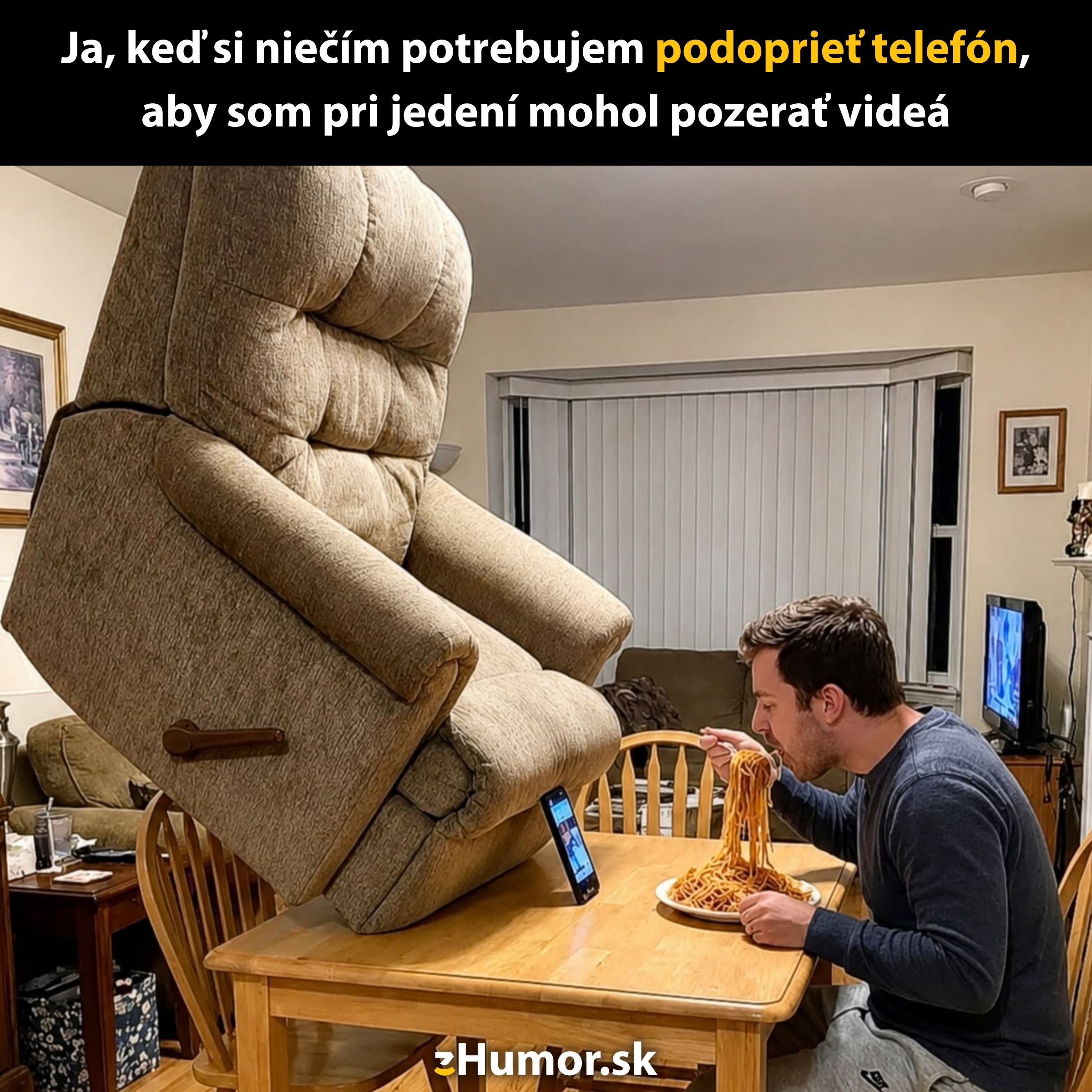 Muž pri jedle sleduje video na mobile, ktorý je podopretý veľkým kreslom na drevenom stole.