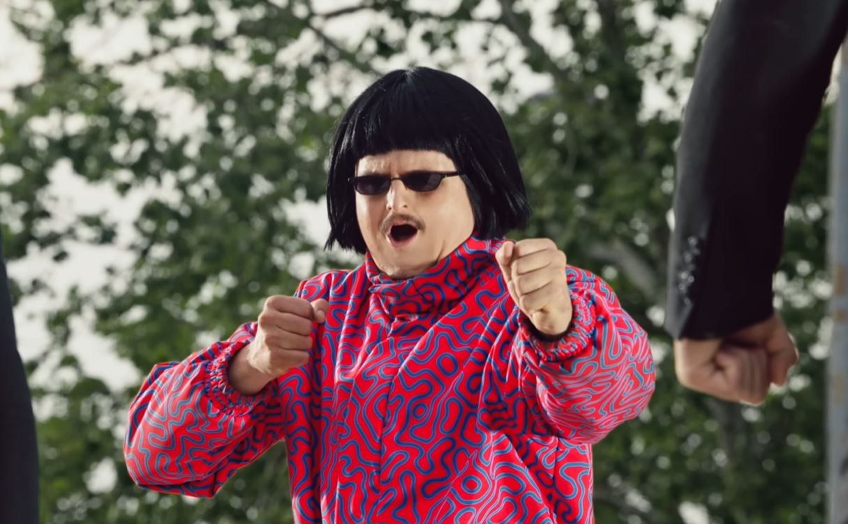 Osoba v pestrofarebnom oblečení s čiernou parochňou predvádza energické pohyby rukami na pozadí zelene.