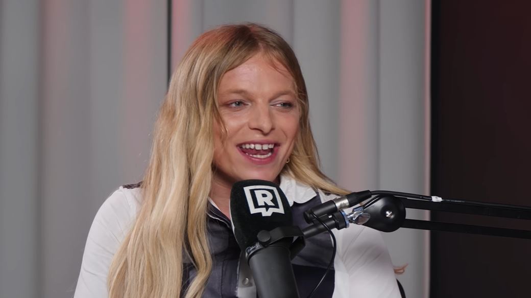Osoba s blond vlasmi hovorí do mikrofónu v štúdiu, biela záclona v pozadí.