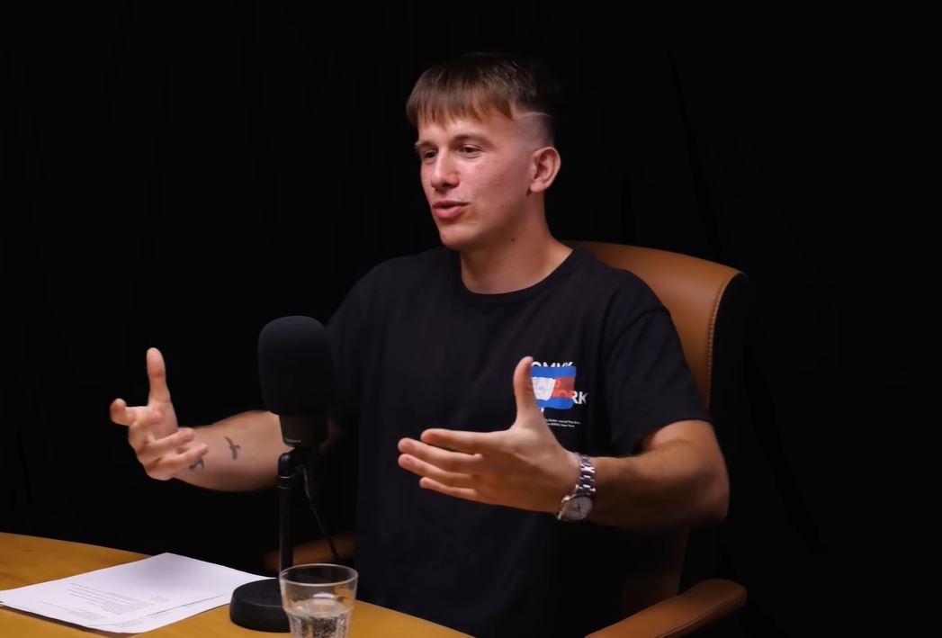 Muž diskusuje do mikrofónu v štúdiu, gestikuluje rukami. Na stole je pohár a papiere.