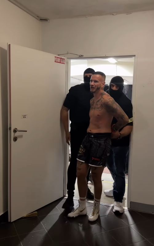 Bojovník v šortkách vychádza z miestnosti, sprevádzaný dvoma mužmi v športovom oblečení.