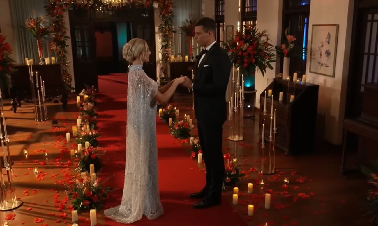 Pár v elegantnom oblečení na červenom koberci, obklopení kvetinovou výzdobou a sviečkami počas romantickej scény.