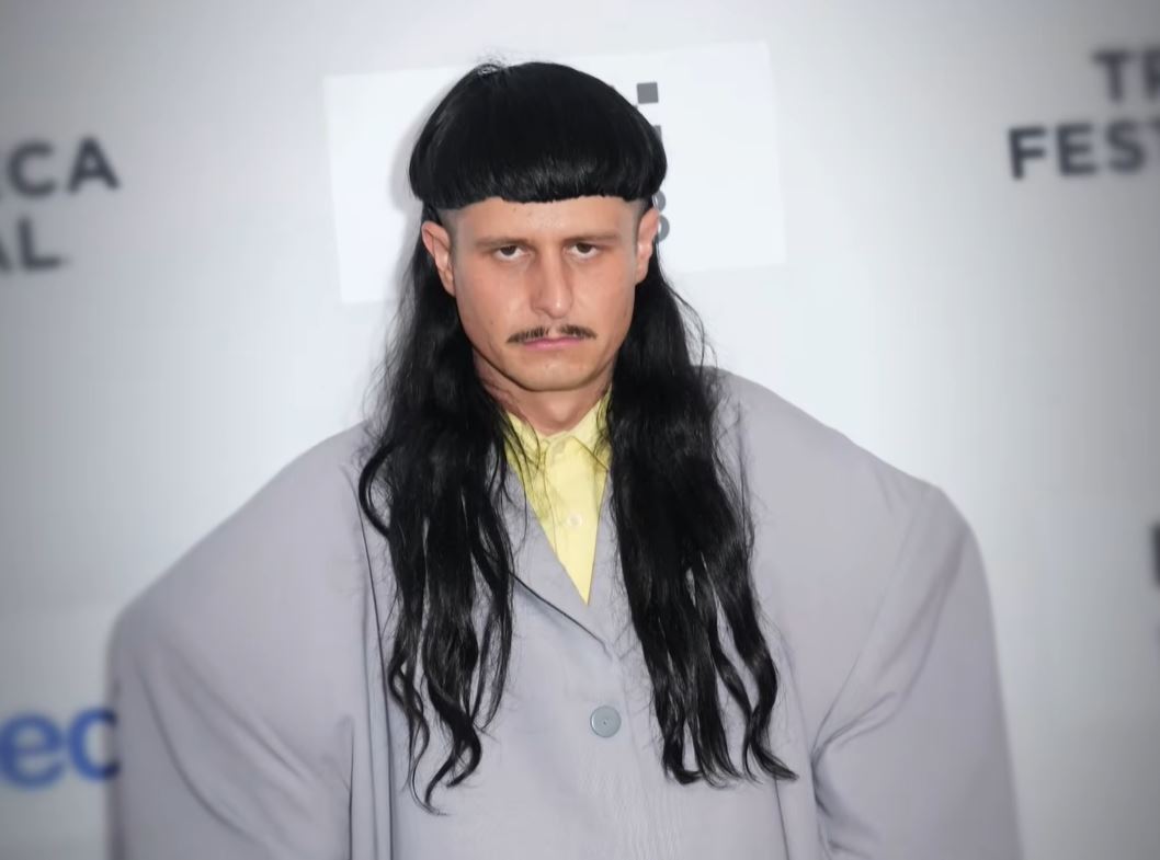 Osoba s dlhými tmavými vlasmi v šedom saku na červenom koberci festivalu Tribeca.