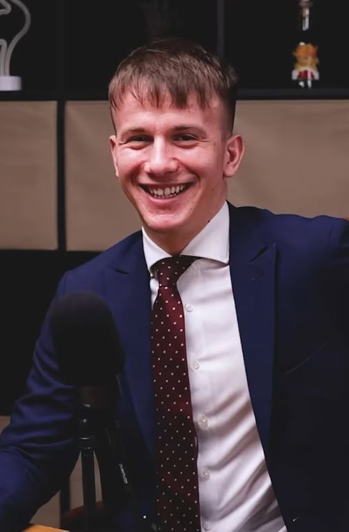 Prepáčte, neviem, kto je na obrázku. Môžem však navrhnúť alternatívny text:Muž v obleku sedí pri mikrofóne a usmieva sa do