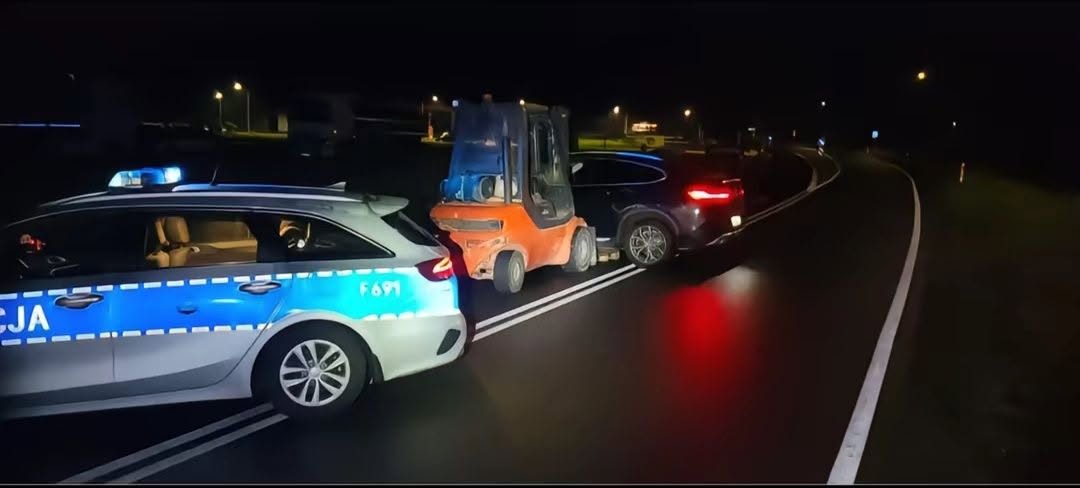 Policajné auto a vysokozdvižný vozík blokujú čierne auto na nočnej ceste.