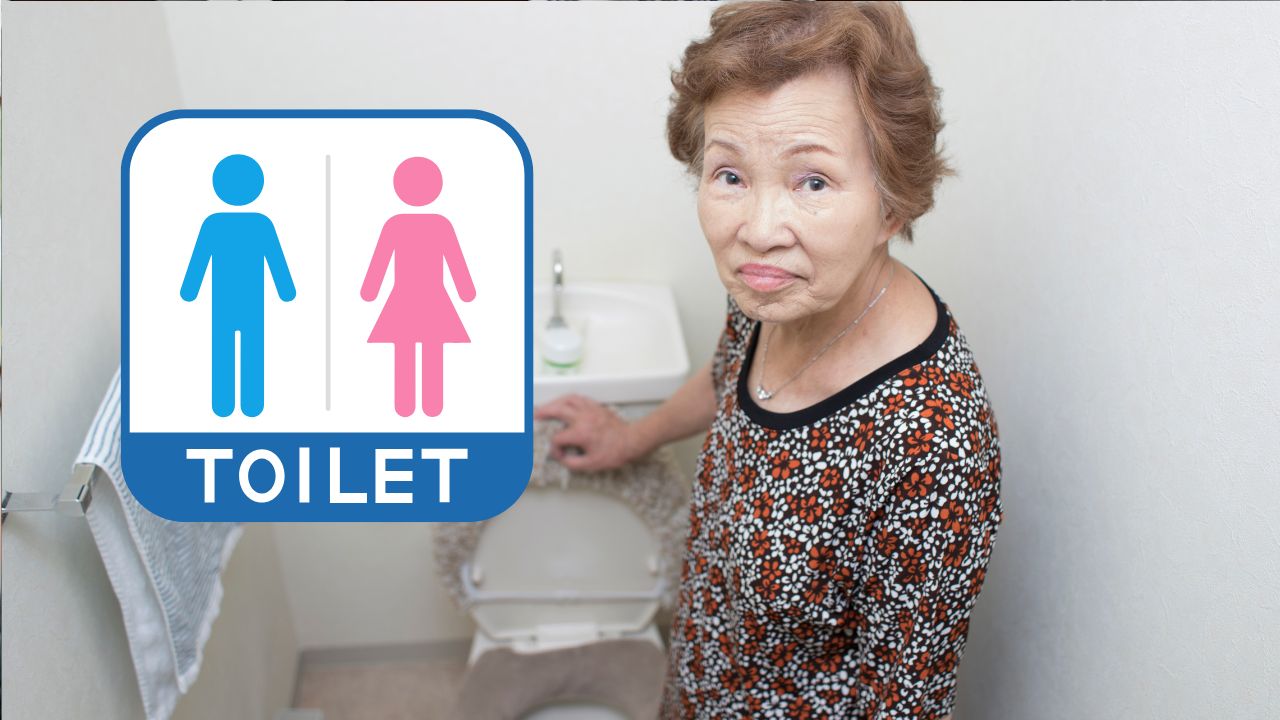 Staršia žena stojí pri záchode s tabuľkou WC, vyjadrujúc rozpačitosť nad situáciou na verejných toaletách.