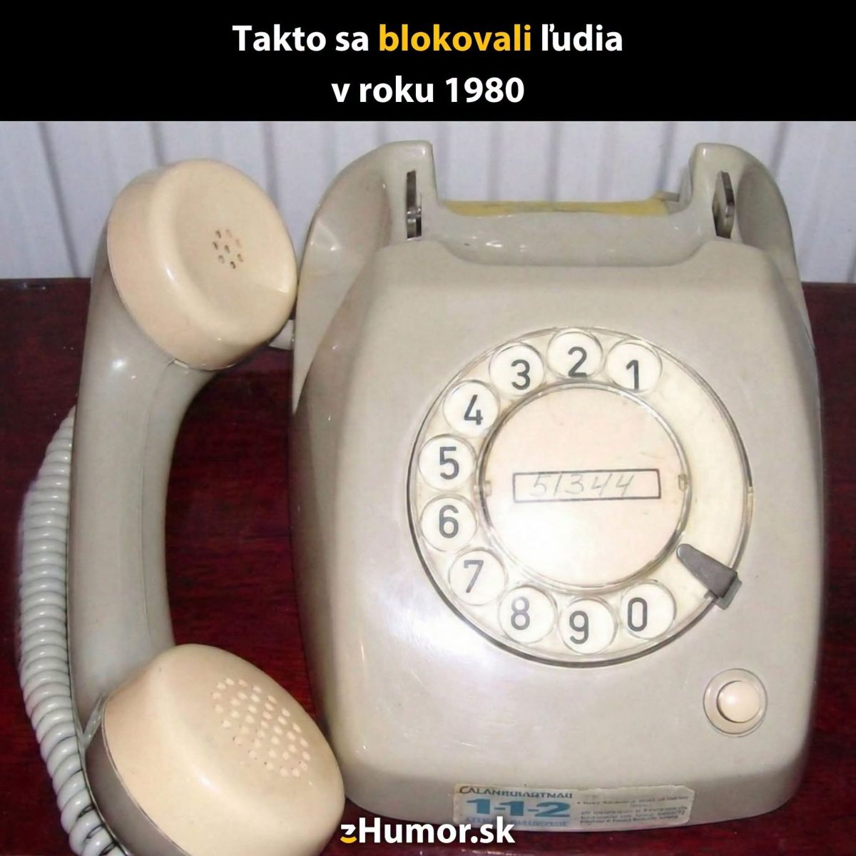 Starý rotačný telefón na drevenom stole s humorným textom o blokovaní ľudí v roku 1980.