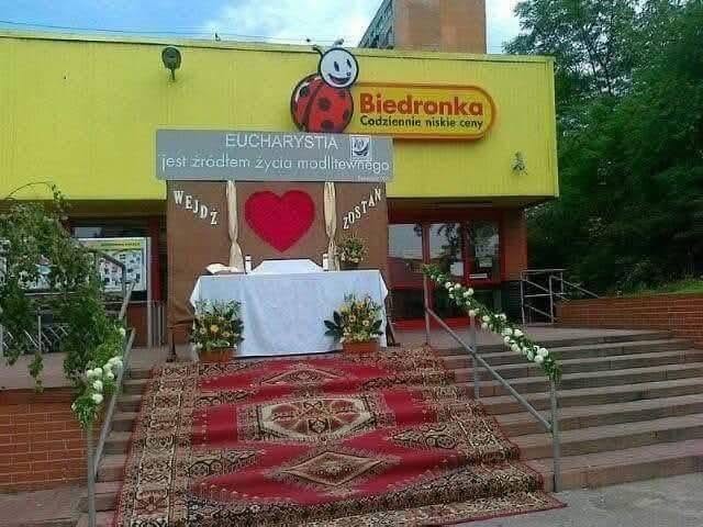 Oltár s kvetmi pred obchodom Biedronka, ozdobený červeným kobercom a s nápismi Wejdź a Zostań.