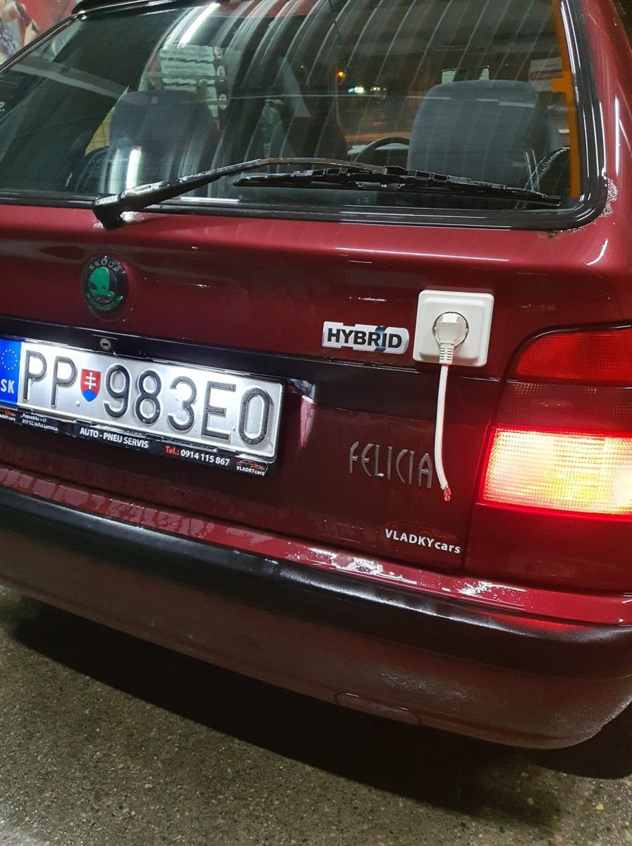 Červené auto Škoda Felicia s falošnou hybridnou nálepkou a humornou zásuvkou na zadnej strane.