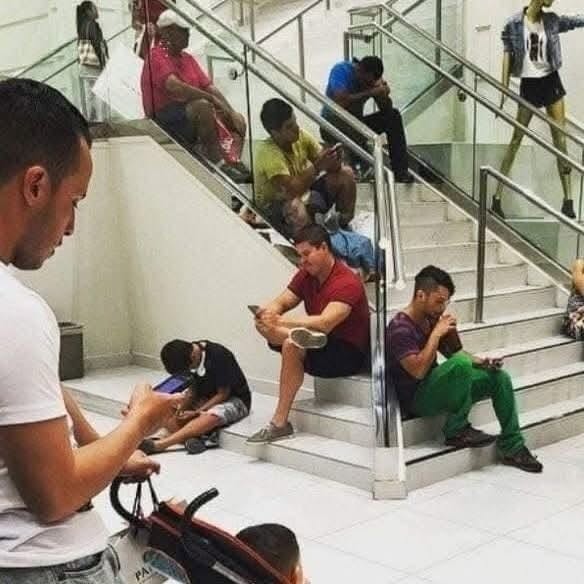 Ľudia v nákupnom centre si krátia čas sledovaním mobilov na schodoch.