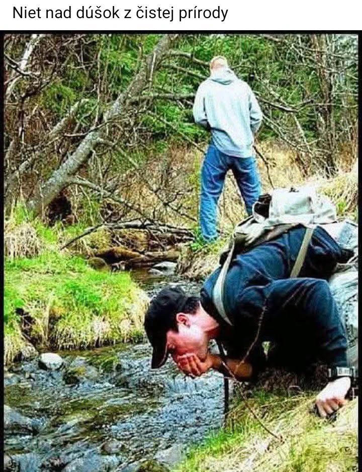 Osoba pije vodu z lesného potoka, zatiaľ čo iný muž stojí v pozadí.