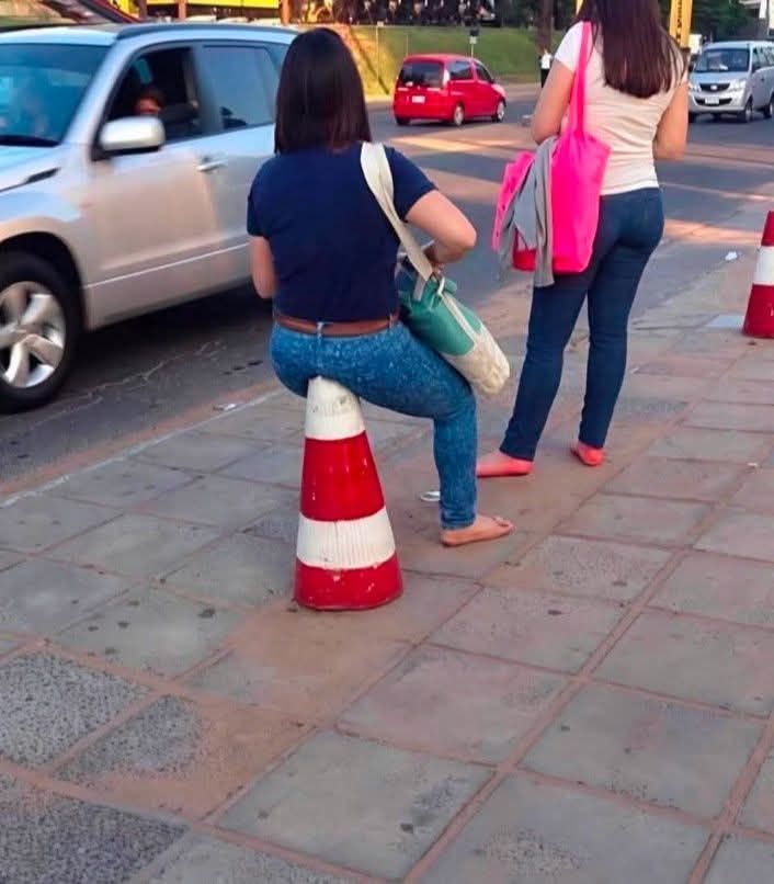 Žena sedí na dopravnej kuželi na chodníku, vedľa prechádzajú autá a kráčajú ľudia.