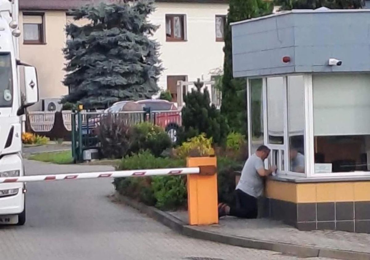 Muž kľačí pri okienku strážnice v areáli pri závore.