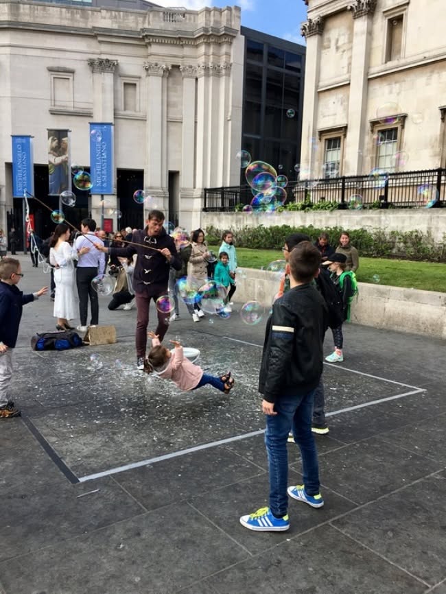 Pouličný umelec tvorí veľké bubliny na námestí, okolo neho sa hrajú deti a sledujú diváci.