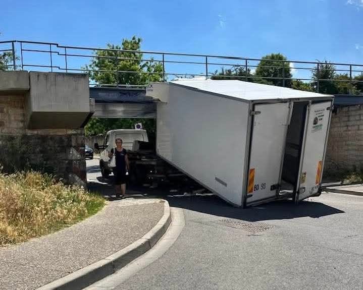 Nákladné auto uviaznuté pod nízkym mostom so zdemolovanou nákladnou časťou na vidiecke ceste.