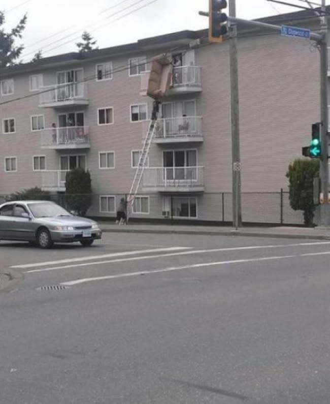 Muž presúva gauč cez balkón na rebríku z ulice k bytu v bytovom dome, zatiaľ čo po ceste prechádza auto.